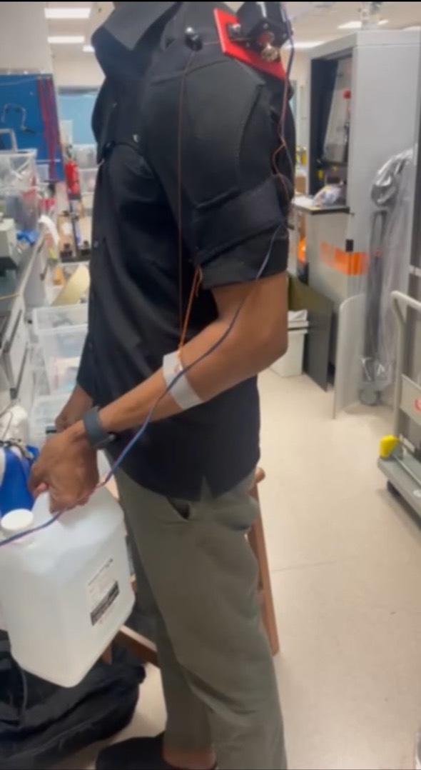 The prototype worn in the lab: shoulder brace with the motor plate, wires down the arm, a forearm band, and a white jerrycan in hand.